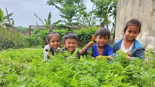 Vier Kinder sitzen lächelnd in einem üppigen grünen Gemüsegarten. Ein Junge zeigt eine frisch geerntete Karotte, die anderen Kinder halten ebenfalls Gemüse in den Händen. Im Hintergrund sind Pflanzen, Bäume und ein Teil einer Mauer zu sehen.