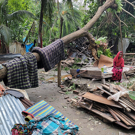 Frau sitzt neben zerstörten Hütten und Häusern umgeben von Wald