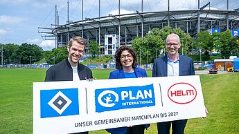 Drei Personen stehen vor dem Stadion und halten ein Banner in der Hand mit den Logos vom HSV, Plan International und der HELM AG