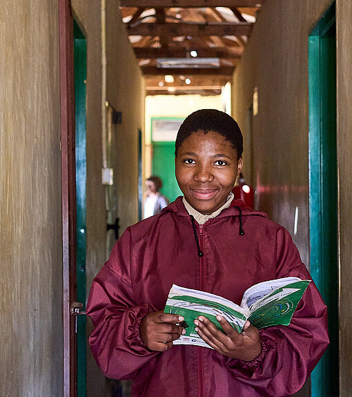 Mädchen in roter Jacke steht im Flur und hat ein aufgeschlagenes Buch in der Hand