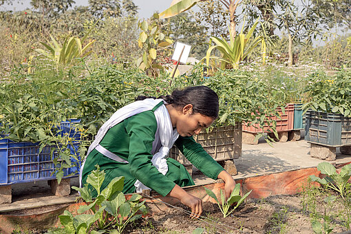 Ein Mädchen in grüner Schuluniform hockt neben einem Hochbeet und prüft die Pflanzen