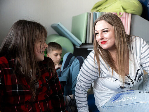 Eine blonde Frau mit roten Lippen sitzt auf dem Boden und lächelt einem Mädchen, das neben ihr sitzt, zu und lehnt sich in ihre Richtung