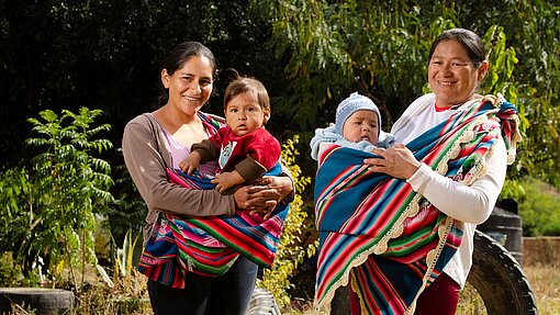 Zwei Frauen stehen nebeneinander im Freien und tragen jeweils ein Baby in bunten Tüchern vor der Brust; im Hintergrund stehen Bäume und große Reifen.