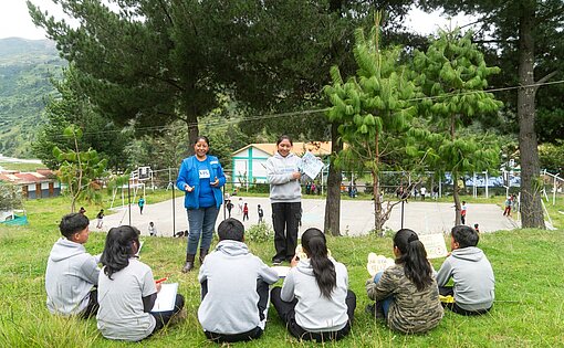 Jugendliche sitzen im Halbkreis auf eine Wiese und hören Lisbeth zu