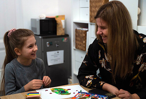 Ein neunjähriges Mädchen sitzt vor selbstgemalten Zeichnungen am Tisch und lächelt ihre Mutter an, die neben ihr sitzt 