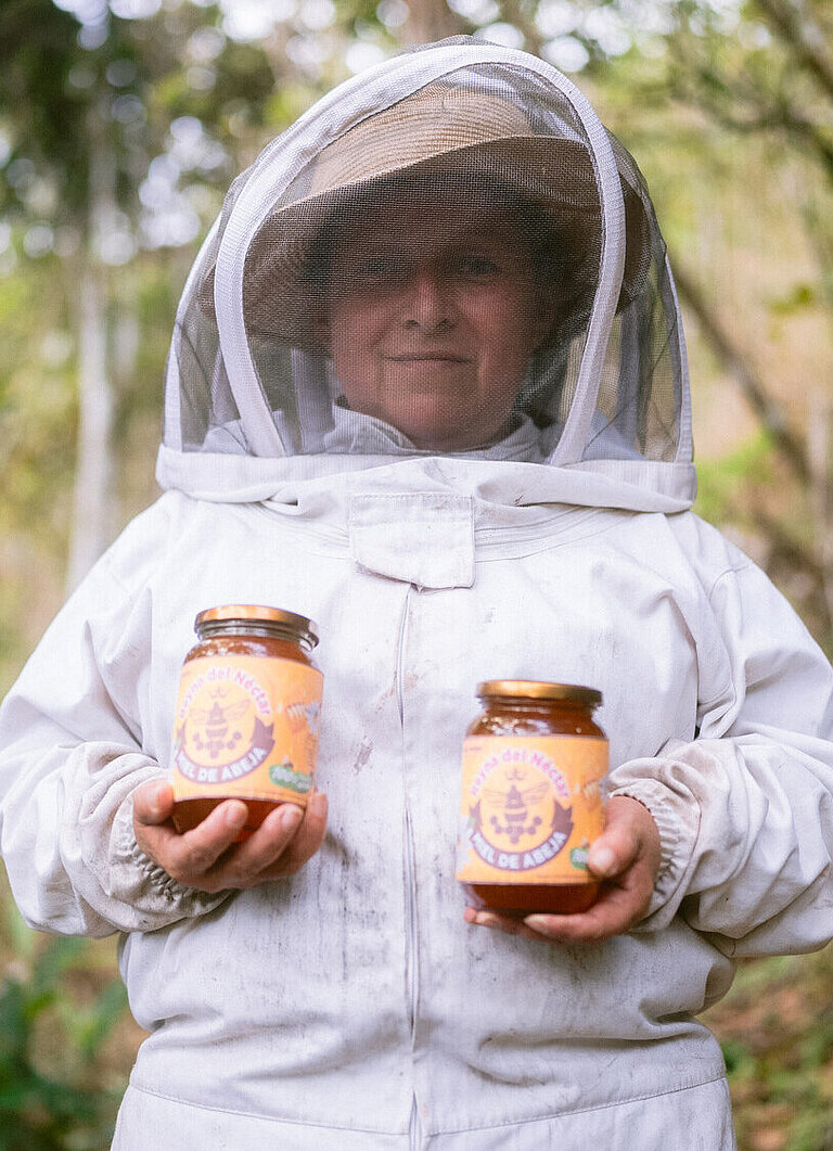 Eine Frau im Imkeranzug steht in einem Wald und hält zwei Honiggläser in den Händen.