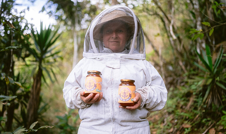 Eine Frau im Imkeranzug steht in einem Wald und hält zwei Honiggläser in den Händen.