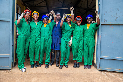 Eine Gruppe junger Frauen in Arbeitsanzügen steht vor einer Werkstatt, sie alle lächeln und haben die Hände in die Luft gehoben