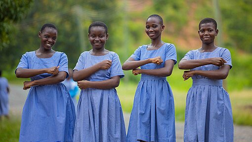 Vier ghanaische Schulmädchen in blaukarierten Schuluniformen stehen nebeneinander im Freien und lächeln in die Kamera. Sie halten ihre Unterarme vor ihrem Körper, sodass sie ein Gleichzeichen bilden.. Die Umgebung ist grün und ländlich.