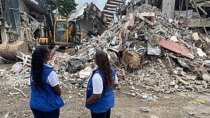 Zwei Frauen mit blauen Plan International Westen stehen vor einem großen Schutthaufen nach einem Gebäudeeinsturz. Eine Frau zeigt auf den Bagger, der im Hintergrund arbeitet.
