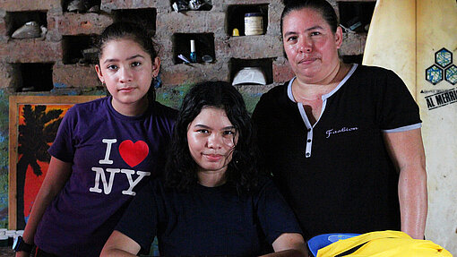 Eine Frau und zwei Mädchen, ihre Töchter, stehen an einem Tisch vor einer Backsteinwand mit kleinen Fächern, in denen verschiedene Gegenstände abgestellt sind. Auf dem Tisch befinden sich bunte Gegenstände und Taschen. Sie schauen mit einem neutralen Gesichtsausdruck in die Kamera.