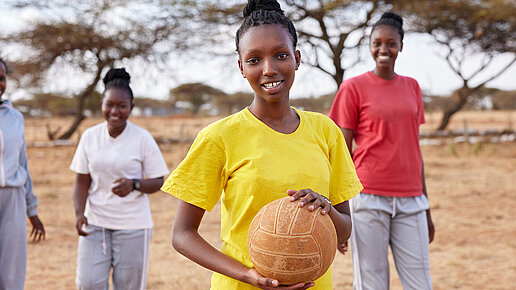 Eine junge Frau trägt ein gelbes T-Shirt und hält einen Ball in der Hand. Hinter ihr stehen ihre Mitspielerinnen.